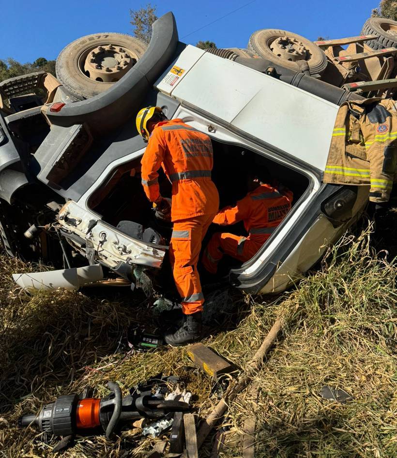 Carreta com madeira tomba em estrada rural e motorista fica preso às ferragens